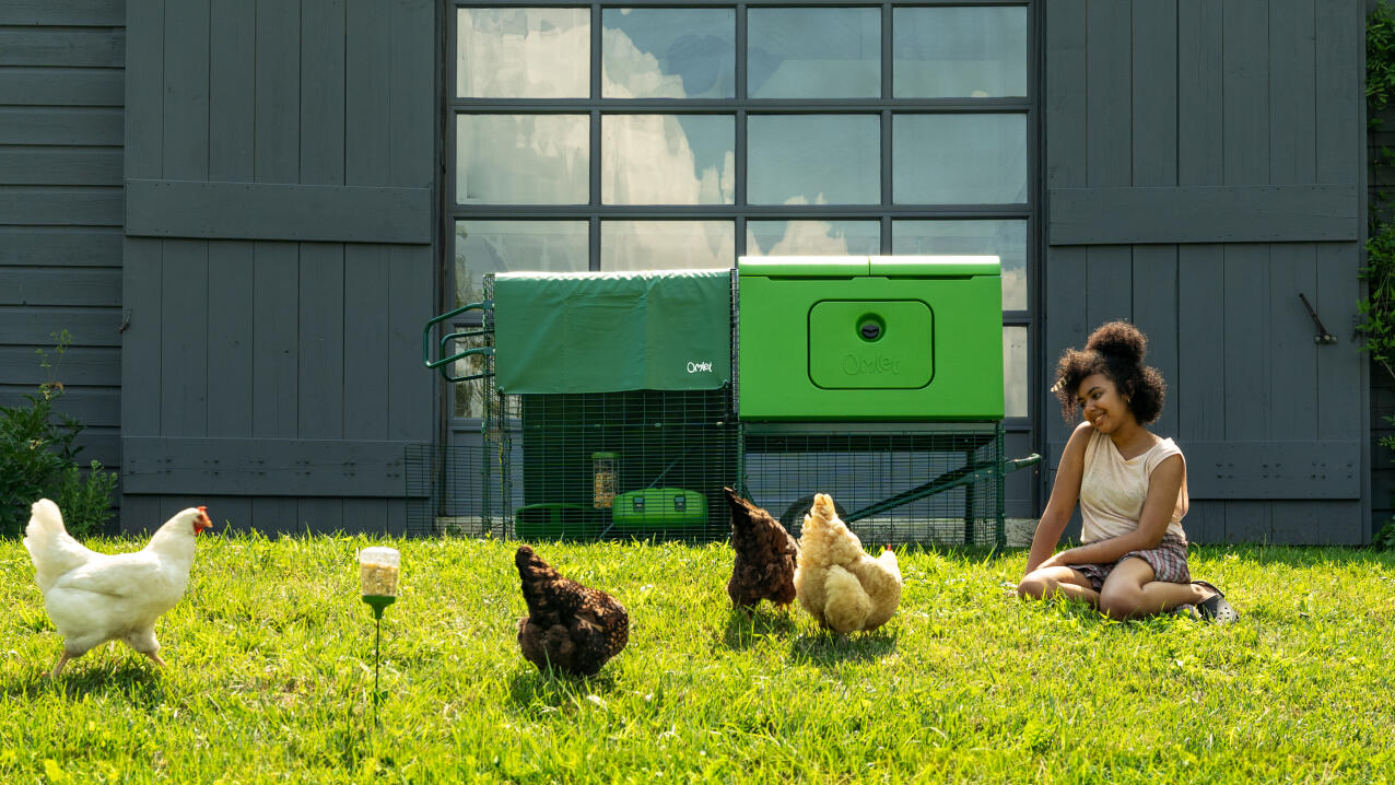 Kobieta siedząca na trawie obok kurnika i obserwująca swoje kury